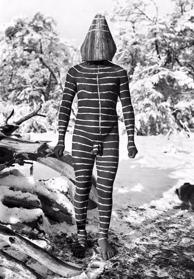 A striped Selk'nam Hain spirit figure standing in snow, in a conical bark mask