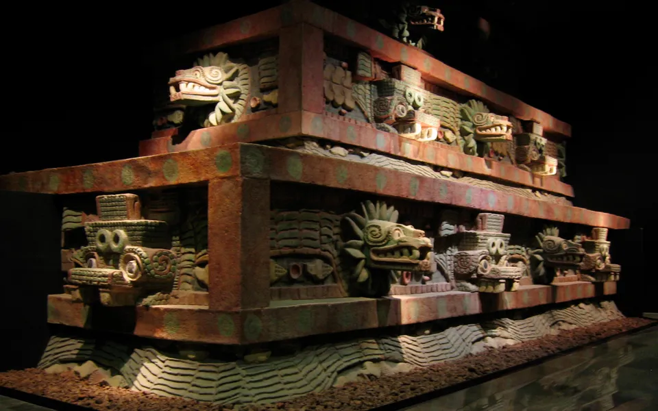 A painted reconstruction of a stepped temple facade lit in a dark gallery: rows of carved feathered serpent heads with bared fangs alternate with blocky goggle-eyed headdresses above a band of carved serpent bodies.