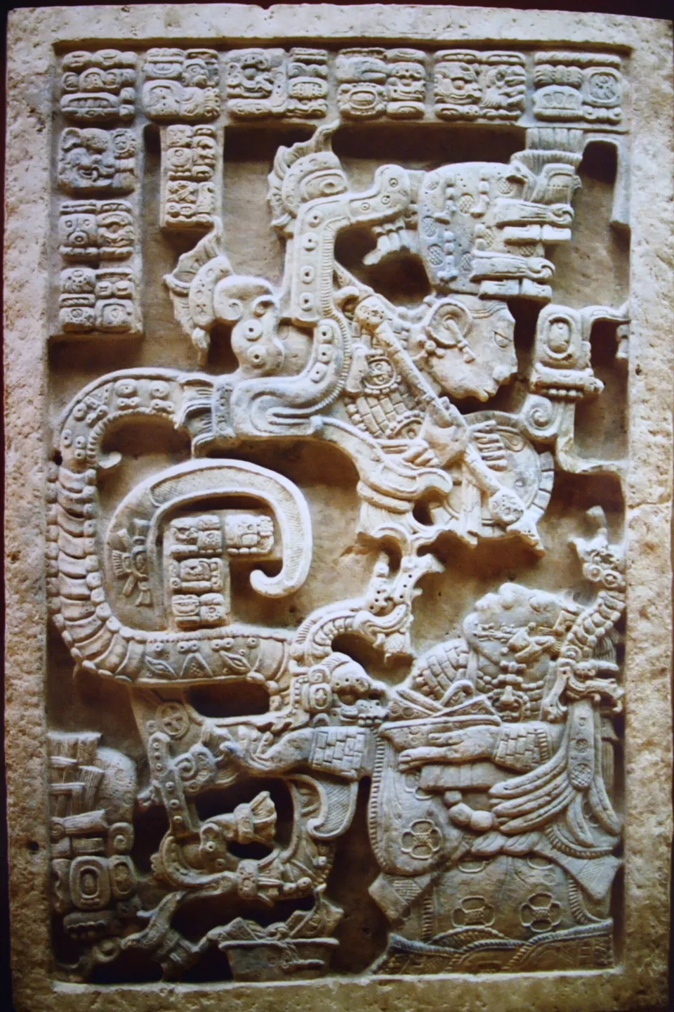 A carved limestone lintel: a kneeling woman holds a bowl at lower right while a great serpent rears up from it, and from the serpent's open jaws a helmeted warrior emerges with spear and shield.