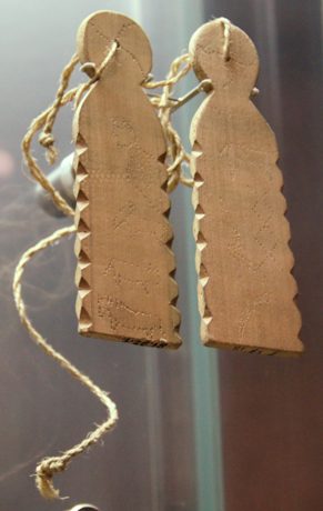 Two pale wooden slats hanging in a display case, their edges cut into saw-teeth, rounded tops pierced for twisted cords, and faces pricked with...