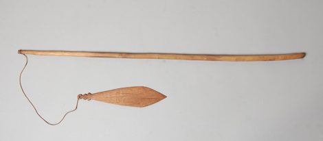 A wooden bull-roarer with a separate carved stick handle, its diamond-shaped blade notched near the cord attachment — shown for the general...