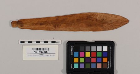 An unfinished bullroarer blank: a rough, leaf-shaped board of pale wood with a pointed tang at one end, photographed with a color card and...
