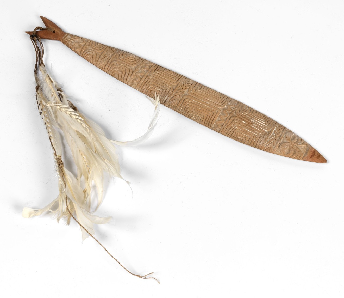Broad carved Marind-region bullroarer decorated with dense incisions and dark pigment, its feathered cord hanging beside it.