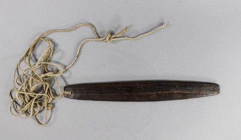 A small dark wooden blade on a long tangled fibre cord, laid on a pale mount beside an old paper museum label reading 'The Voice of Oro.'