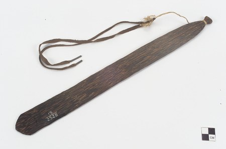 The Miskito bull-roarer itself — an elongated palm-wood blade with its hide-lace cord wrapped at one tip, catalog number 13/2528 inked on the...