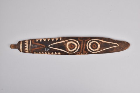A second Maipua blade, collected by J. H. Holmes in the 1890s: paired circular motifs inside pointed outlines, bordered by pale triangles and bands, with a short wooden tab at one end.