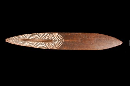 A wooden rhombe carved along one half with chevron zigzags rising into a stylized face, with a small perforation at its pointed tip — the...