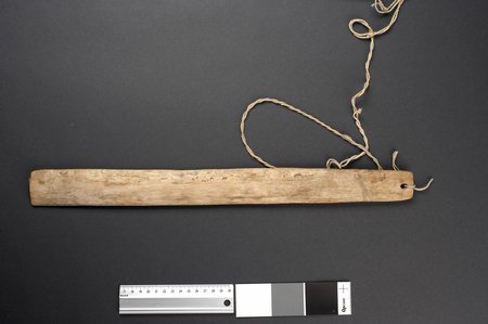 A narrow, nearly rectangular pale board with blunt ends, its weathered face streaked with dark mottling, a twisted cord tied through the hole...