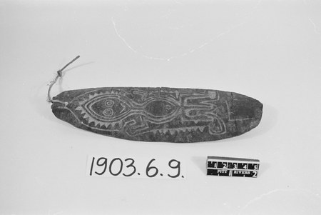 A bark or hard-wood blade carved with a stylised ancestor face — ringed eyes above a toothed, triangular border — its cord knotted through the...