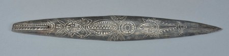 A dark carved wooden blade from the Huon Gulf, its surface divided into bands of chevrons, faces and circular motifs: a bullroarer collected by...