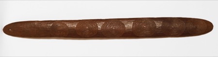 A long, narrow dark wooden blade with adze-marked surface, six carved spiral roundels along its length and a small hole near the left end.