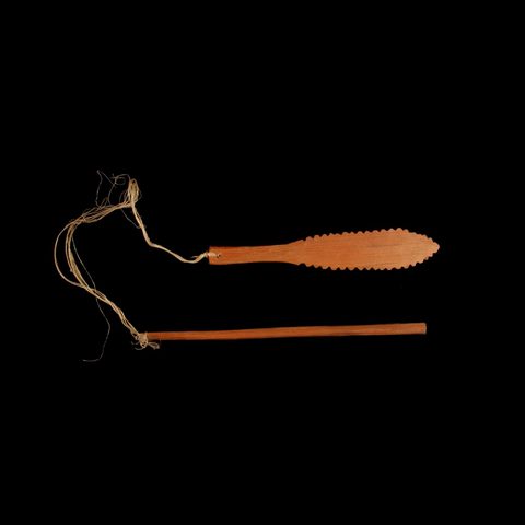A carved wooden blade with a serrated, feather-like edge on one end and a plain handle on the other, lying beside its separate cord-wound wooden grip stick.