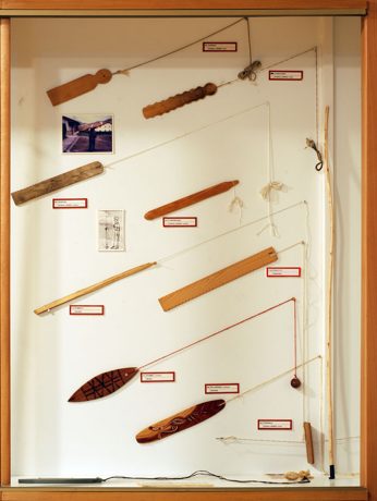 A glass display case of free aerophones at Soinuenea, with a plain wooden burruna board and its cord visible among rombos and other whirled...