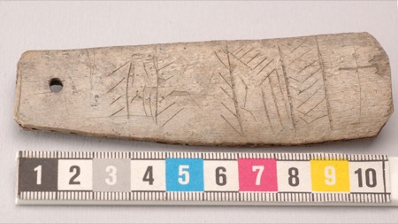 A small grey bone blade, tapering slightly, with a round hole near its left end and bands of incised chevrons, above a 10 cm colour scale.
