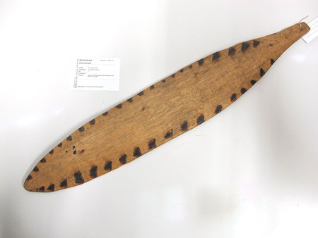 A large flat wooden Schwirrholz with a line of small burnt-black notches cut along both edges, tapering to a point at the swinging end....