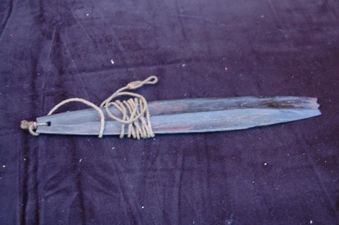 A long dark wooden bullroarer blade on dark cloth, its cord wound round the blade near the pierced end.