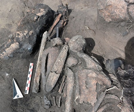 An excavation photograph shows a long bone blade in two pieces lying over a clay figure among dark grave deposits, beside a scale and north arrow.