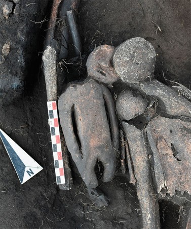 The clay figure lies in the grave with its round detached head beside its shoulder, next to human skeletal remains and an excavation scale.
