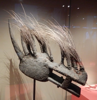 A Bamana Komo mask from Mali, its mud-blackened wood bristling with long fibre quills between two curving horns — a glimpse of the...