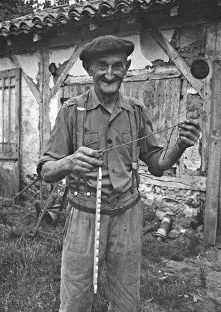 A man in a beret holding up a notched wooden bullroarer by its cord outside a building.