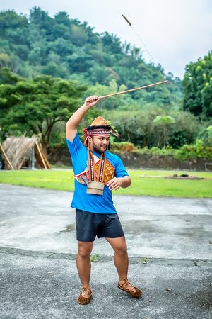 A man in Tsou dress and a feathered headband raises a long rod as a euvuvu blade swings from a cord at its tip.