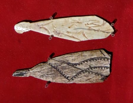 Two engraved Magdalenian bone blades arranged one above the other on a red background; the upper is finely incised and the lower has toothed zigzag bands.