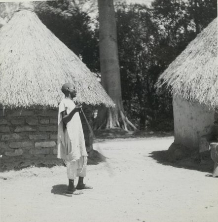 Black-and-white photograph of a Kissi man in a light robe, standing in profile between two round thatched houses at Kenéma Pombo, Guinea, in 1947, holding a cord descending beside him.