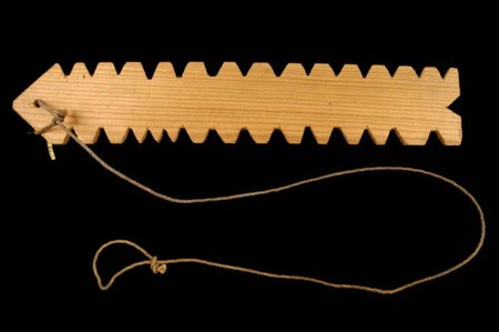 A wooden rhombe made at Commensacq, with V-shaped notches along both long edges and a looped cord at one end.