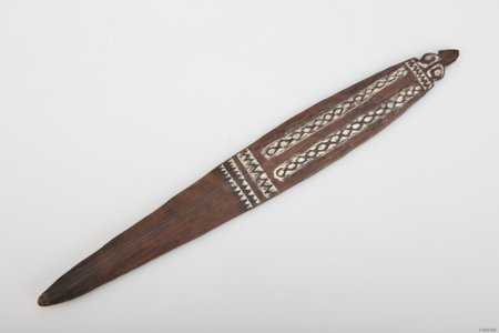 A long tapering dark wooden bullroarer with white-filled bands of looping and zigzag carving near its pointed upper end.