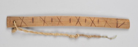 A wooden board burned with alternating X and single-stroke marks down its length, cord tied through a hole at one end: an African bullroarer of...