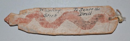A board inscribed in ink "Whirling Stick — To Scare the Devil" beside a painted red serpentine line — a North American bull-roarer held by the...