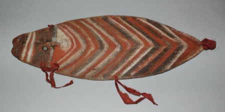 A painted New Guinea bull-roarer board with a carved face at one end, shell-inlay eyes set above red, white, and black chevron banding, and red...