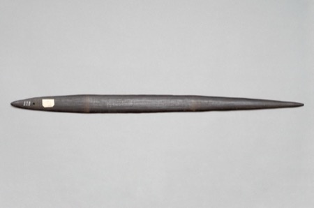 A dark, double-pointed wooden blade, largely unadorned but for its painted accession number, a torn scrap of paper label, and the name...