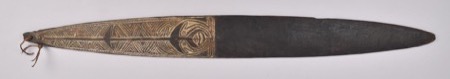 A dark wooden blade carved in white-filled relief with a stylized bird-like face - a generic New Guinea bullroarer, not the urubaia this page...