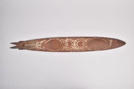 A wooden bullroarer carved in relief with mirrored spiral and face motifs framed by triangulated bands, its notched tip pierced for the cord -...