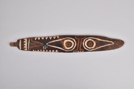 A dark wooden board painted with a white triangular tooth border and two large eye-like circles, its narrow tang notched — a New Guinea...