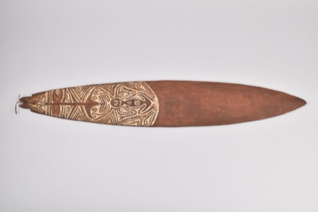A New Guinea bull-roarer blade half covered in white-incised chevron banding around a stylized bird-headed figure, the remaining half left...