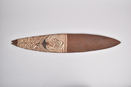 A wooden board painted with pale zigzag bands framing a stylized face, its butt carved into small serrated notches — a New Guinea bull-roarer...