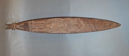 A deeply incised New Guinea bull-roarer showing a stylized ancestor figure with large eyes and articulated limbs, its cord looped through the...