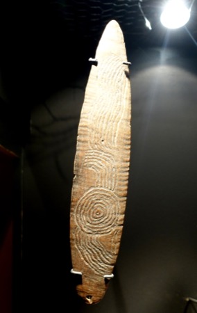 A Central Australian tjurunga board, its surface deeply incised with concentric circles and wavy parallel lines — shown for the general...