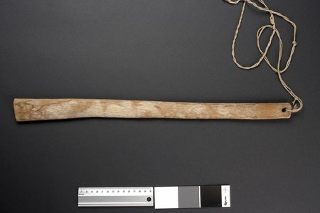 A pale, faintly bowed wooden slat with a cord threaded through a hole at one end - one of a set of Hopi bullroarers in Oslo's ethnographic...