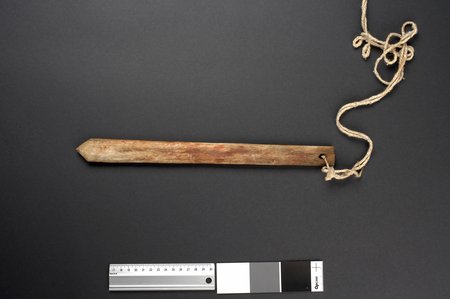 A pale wooden slat blotched with reddish stain, smooth-edged, cut to a point at one end and squared at the other where a frayed cord passes...