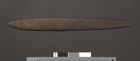 A long dark bull-roarer blade, carved only near one tip — a small notched tang, zigzag-and-triangle bands, and a slim central column between...