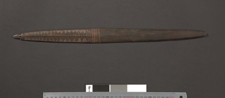 A long dark New Guinea bull-roarer, rows of incised triangles running down the first third of the blade to a red-banded zigzag, the rest left...