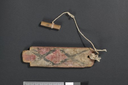 A small rectangular wooden bullroarer painted with a bold pink-and-black diamond-and-cross design, its cord ending in a short wooden toggle...