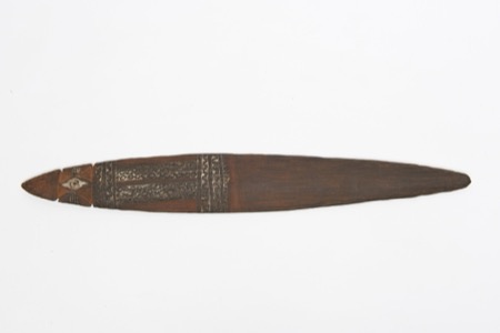 A dark, carved New Guinea bull-roarer, its head end bearing a white-rimmed eye between notched jaws and a broad band of chain-like interlocking...