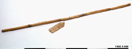 A long, thin cane rod wrapped with cord and hung with a small wooden tag - a Pipil bullroarer from the same Stockholm collection, standing in...