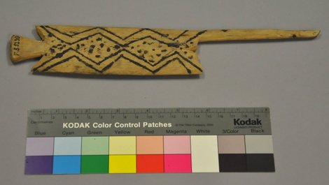 A carved wooden blade ending in a small splayed peg at one tip and a long slender shaft at the other, its face painted with a dotted...