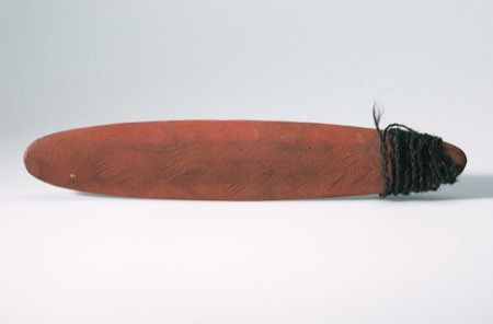 A Luritja bullroarer worked in red ochre, one end carved with a spiral, the rest banded with crossing wavy lines, and dark cord wound at the...