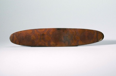 A red-ochred Aboriginal Australian bullroarer, its face marked with three clusters of concentric circles — shown for the general Central...